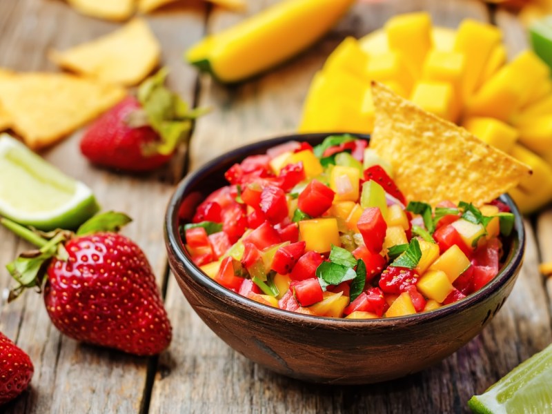 Eine Schale Erdbeer-Mango-Salsa mit einem Nacho, drumherum Erdbeeren, Mango und Limettenspalten.