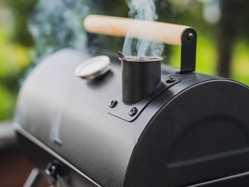 Ein geschlossener Grill, der aus einem kleinen Schornstein heraus raucht. Im Hintergrund kann man verschwommen eine Wiese erkennen.