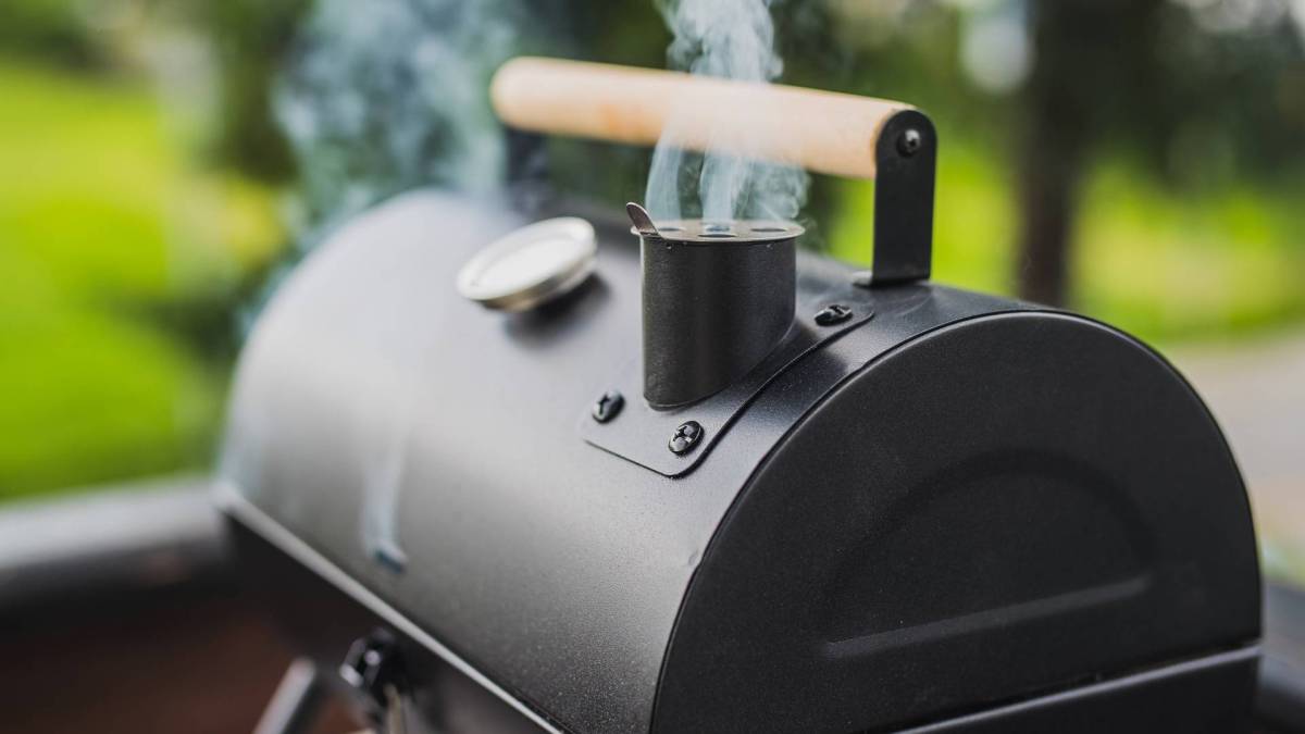 Ein geschlossener Grill, der aus einem kleinen Schornstein heraus raucht. Im Hintergrund kann man verschwommen eine Wiese erkennen.