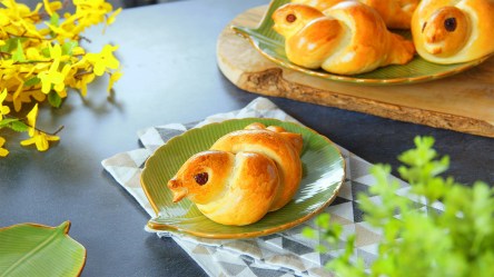 Eine Hefezopftaube liegt auf einem grünen, blattförmigen Teller. Im Hintergrund liegen weitere Hefezopftauben. Der Teller steht auf einem Küchentuch. Im Vordergrund sowie im Hintergrund ist etwas Pflanzendeko.