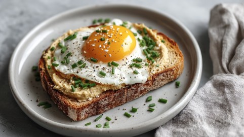 Ein Hummus-Spiegelei-Toast mit Schnittlauch bestreut auf einem Teller, daneben eine Stoffserviette.