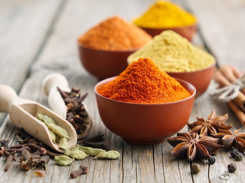 Ein paar Schalen mit Gewürzen. Außerdem liegt einzelner Sternani sowie Nelken und Kardamomkapsel gemeinsam mit zwei kleinen Gewürzschaufeln neben den Schalen verteilt. Diese Gewürze werden verwendet, um indische Gewürzmischungen herzustellen.