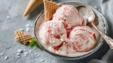 Eine Schüssel mit drei Kugeln Joghurt-Rhabarber-Eis. Außerdem steckt eine Waffel im Eis und Eiswaffeln liegen neben der Schüssel. In der Schüssel ist auch Löffel.