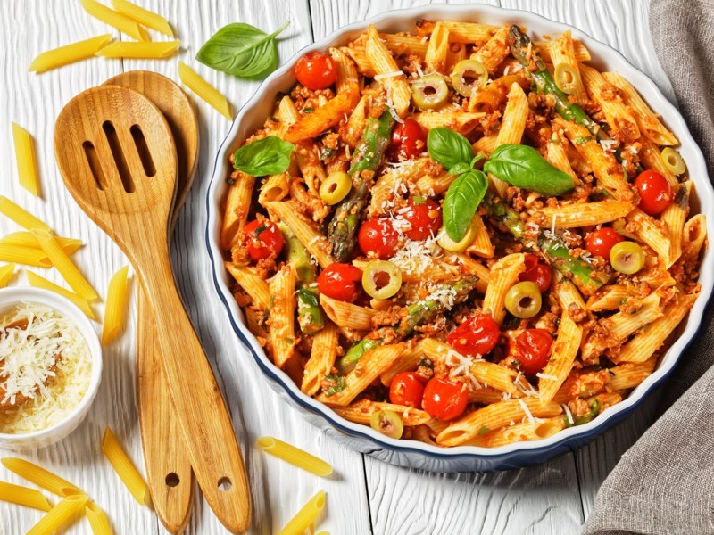 Nudelauflauf mit Spargel und Hack in einer runden Auflaufform. Daneben liegen Holzbesteck und Nudeln.