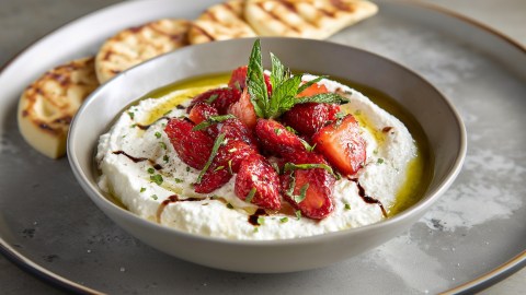 Eine Schale mit Ricotta-Dip mit gerösteten Erdbeeren. Die Schale steht auf einem großen Teller. Im Hintergrund kann man drei kleine Pita-Brote erkennen.