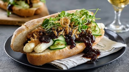 Spargel-Hotdog mit dunkler Soße, Salat und Gurkenscheiben auf einem Teller, im Hintergrund ein Glas und ein weiteres Hotdog.