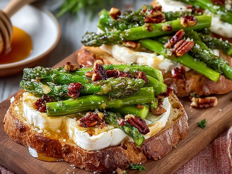 Ein Holzbrett mit mehreren Scheiben Spargel-Toast mit Camembert, die mit Honig und Nüssen garniert sind. Im Hintergrund kann man einen Teller mit einem Honiglöffel aus Holz und Honig erkennen.