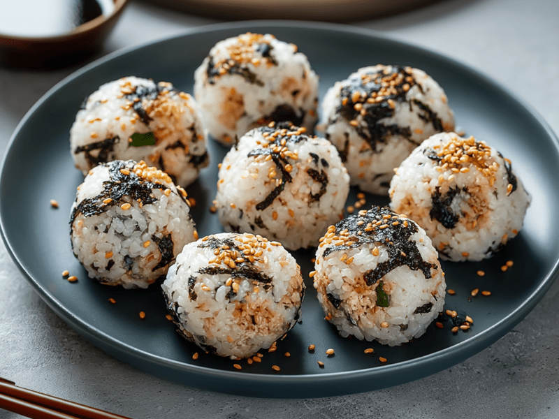 Ein Teller mit einigen Thunfisch-Reisbällchen. Außerdem sind im Bild ein Paar Stäbchen zu sehen und eine Schale mit Sojasoße.