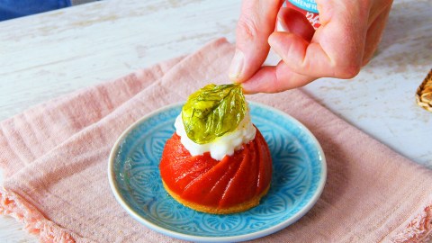 Ein Teller mit Tomaten-Panna-cotta, die mit einem Mozzarella-Schaum und einem frittierten Basilikumblatt garniert ist. Man sieht, wie eine Hand, das Basilikumblatt auf den Mozzarella-Schaum setzt. Der Teller steht auf einem rosafarbenen Tuch.