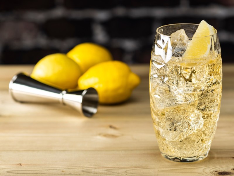 Ein Glas mit Whisky Soda steht auf einem Holztisch. Im Hintergrund liegen drei Zitronen sowie ein Cocktail-Messbecher.