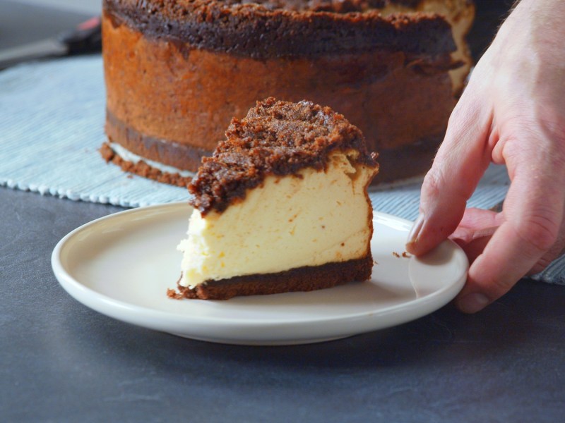 Ein Teller mit einem Stück Zimtschnecken-Käsekuchen. Außerdem ist eine Hand zu sehen, die den Teller gerade hinstellt. Im Hintergrund ist der restliche Kuchen zu sehen.