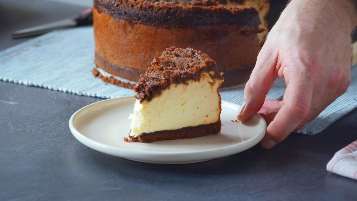 Ein Teller mit einem Stück Zimtschnecken-Käsekuchen. Außerdem ist eine Hand zu sehen, die den Teller gerade hinstellt. Im Hintergrund ist der restliche Kuchen zu sehen.
