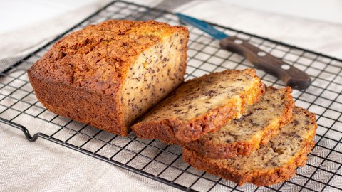 Ein angeschnittenes Zitrone-Bananenbrot mit Mohn auf einem Kuchengitter, daneben ein Messer.