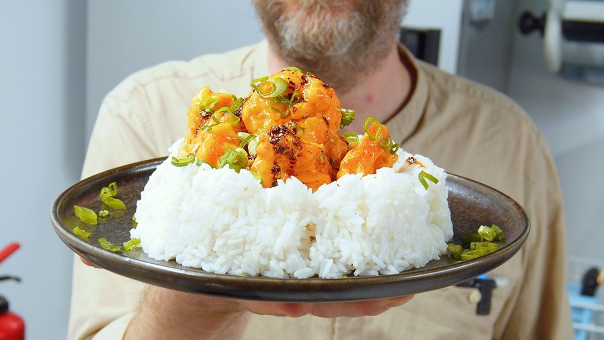 Ein Teller mit einer großen Portion Reis, auf der die Bang Bang Garnelen verteilt sind. Koch Oli hält den Teller in die Kamera.