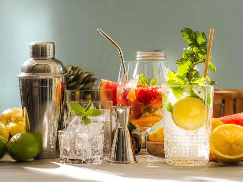 Einige Gläser mit erfrischenden Drinks, ein Cocktailshaker sowie ein Glasgefäß mit Eis. Außerdem sind sehr viele frische Früchte um die Gefäße und Gläser verteilt.