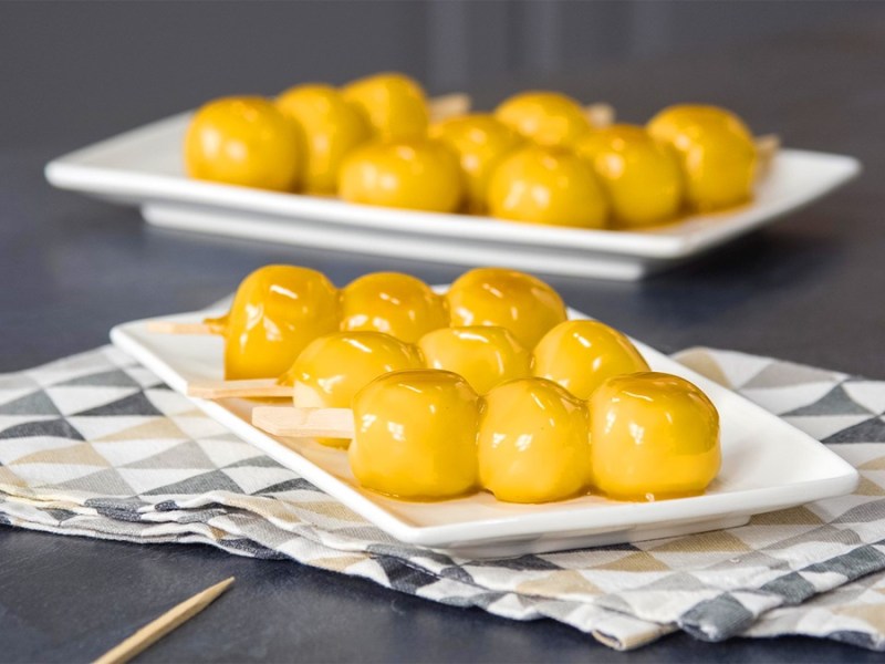 Drei Dango-Spieße liegen auf einem weißen Teller. Im Hintergrund sieht man weitere Dango-Spieße. Sie sind mit einer goldfarbenen Glasur übergossen.