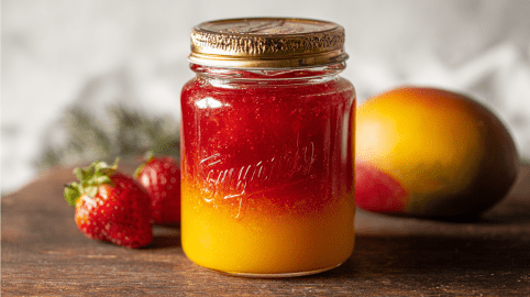 Ein Glas Erdbeer-Mango-Marmelade. Im Hintergrund zwei Erdbeeren und eine Mango.
