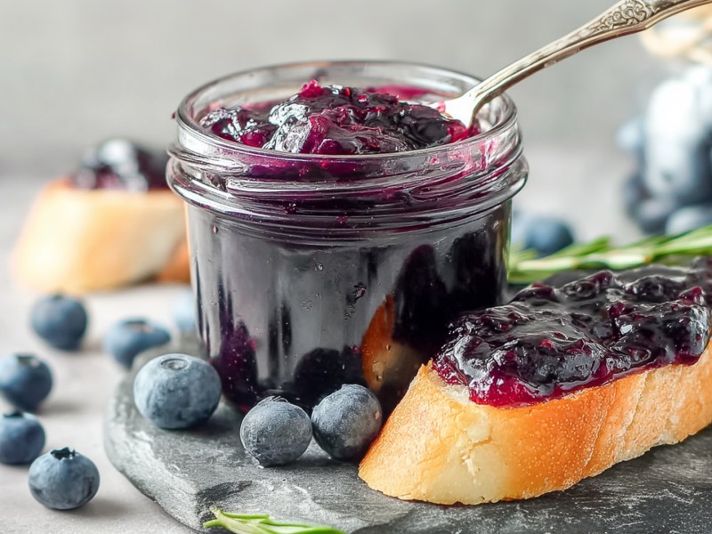 Ein Einmachglas mit herzhafter Blaubeer-Zwiebel-Marmelade, in der ein Löffel steckt. Neben dem Glas liegt eine Scheibe Baguette, die mit der Marmelade bestrichen ist. Ringsum liegen einzelne Blaubeeren herum.