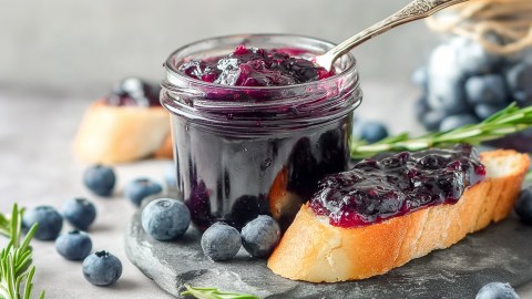 Ein Einmachglas mit herzhafter Blaubeer-Zwiebel-Marmelade, in der ein Löffel steckt. Neben dem Glas liegt eine Scheibe Baguette, die mit der Marmelade bestrichen ist. Ringsum liegen einzelne Blaubeeren herum.