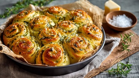 Eine runde Backform mit herzhaften Kräuterschnecken. Die Backform steht auf einem Holzbrett, dass mit einem Küchenhandtuch dekoriert ist. Ringsum verteilt liegen frische Kräuter. Außerdem ist ein Stück Parmesan sowie eine kleine Schale mit grobem Meersalz zu sehen.