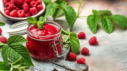 Ein Glas Himbeermarmelade, dahinter eine Schale mit frischen Himbeeren. Drum herum liegen einige Himbeerblätter.