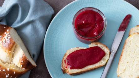 Teller mit zwei Scheiben Brot, bestrichen mit Himbeermarmelade ohne Kerne, Messer und ein Glas Marmelade daneben
