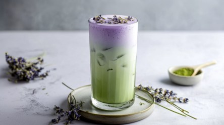 Ein Glas mit Iced-Lavendel-Matcha-Latte, das auf einem Keramikuntersetzer steht. Ringsum liegen einige Zweige Lavendel herum. Außerdem steht eine kleine Schale mit Matcha-Pulver im Hintergrund.