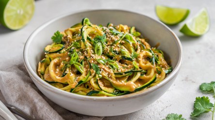 Eine große Schüssel voll kalter Zucchini-Nudeln mit Erdnuss-Sesam-Soße, die mit Sesam sowie Frühlingszwiebeln garniert sind. Um die Schüssel herum liegen Limettenschnitze sowie etwas frischer Koriander. Auch ein Küchenhandtuch ist zu sehen.