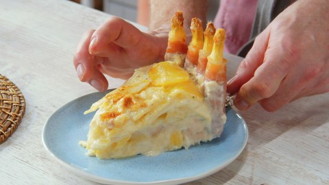 Ein Stück Kartoffel-Spargelauflauf auf einem Teller. Es sind die Hände des Kochs zu sehen.