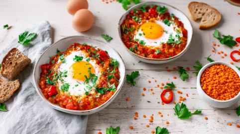 Zwei Schalen mit Linsen-Paprika-Schakshuka steben nebeneinander. LInsen, Kräuter und Eier liegen als Deko daneben.