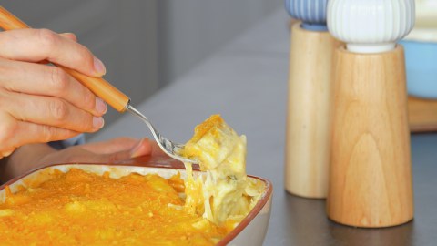 Eine Auflaufform mit Maultaschen-Auflauf mit Spargel, die auf einer Arbeitsfläche steht. Man kann erkennen, wie eine Hand mit einer Gabel etwas aus dem Auslauf herausgabelt. Neben der Form kann man einen Salz- und Pfefferstreuer erkennen.