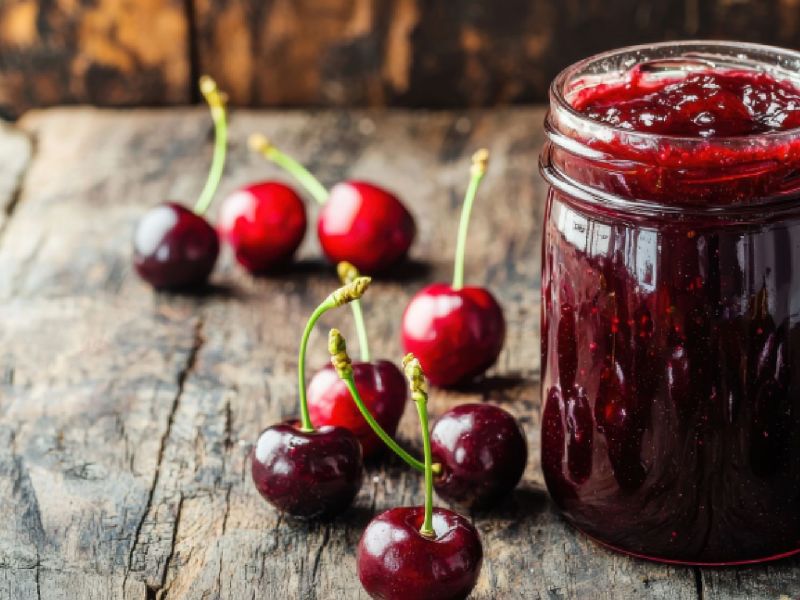 Ein Glas Sauerkirschmarmelade, im Hintergrund und daneben liegen Sauerkirschen.