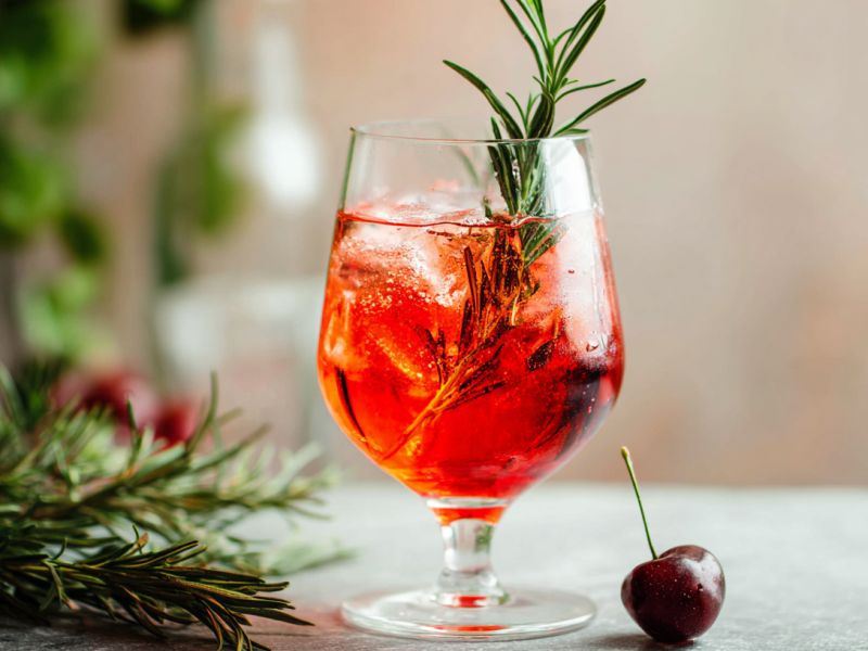 Ein Glas Savoia Spritz, garniert mit Rosmarin und einer Kirsche.