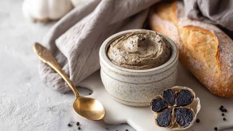 Schale mit schwarzem Knoblauch, eine aufgeschnittene Knolle und Baguette daneben