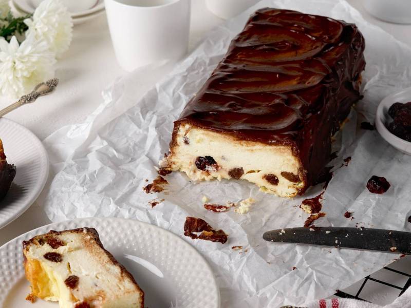 Ein weißes Backpapier auf dem ein angeschnittener ukrainischer Käsekuchen liegt. Auf Tellern, die daneben stehen, liegen Stücke des Kuchens. Außerdem kann man ein paar weiße Blumen erkennen und es steht eine Schale mit Rosinen da. Ein Messer liegt beim Kuchen.