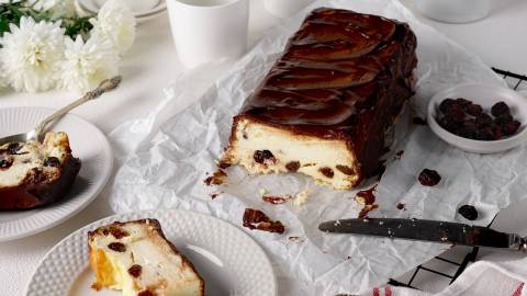 Ein weißes Backpapier auf dem ein angeschnittener ukrainischer Käsekuchen liegt. Auf Tellern, die daneben stehen, liegen Stücke des Kuchens. Außerdem kann man ein paar weiße Blumen erkennen und es steht eine Schale mit Rosinen da. Ein Messer liegt beim Kuchen.