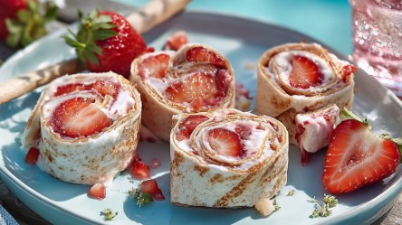 Vier süße Wrap-Röllchen mit Erdbeeren auf einem Teller. Drumherum liegen frische Erdbeeren.