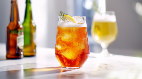 Ein Glas Aperol-Bier-Spritz steht auf einer Marmor-Anrichte. Links im Hintergrund stehen zwei Bierflaschen, rechts im Hintergrund ein Bierglas.