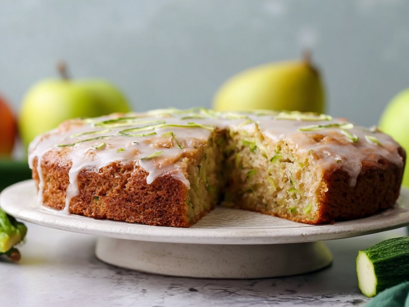 Ein runder Apfel-Zucchini-Kuchen, der auf einer Kuchen-Etagere liegt. Ringsum liegen frische Äpfel und Zucchini verteilt.