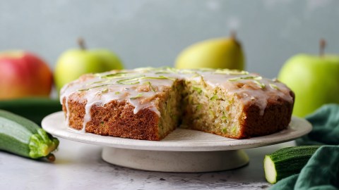 Ein runder Apfel-Zucchini-Kuchen, der auf einer Kuchen-Etagere liegt. Ringsum liegen frische Äpfel und Zucchini verteilt.