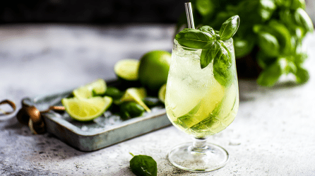 Ein Glas Basilikum Spritz vor einem Tablett mit Limetten und etwas Basilikum.