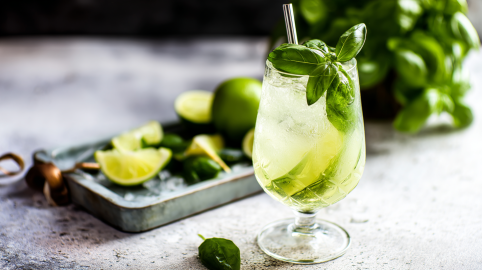 Ein Glas Basilikum Spritz vor einem Tablett mit Limetten und etwas Basilikum.