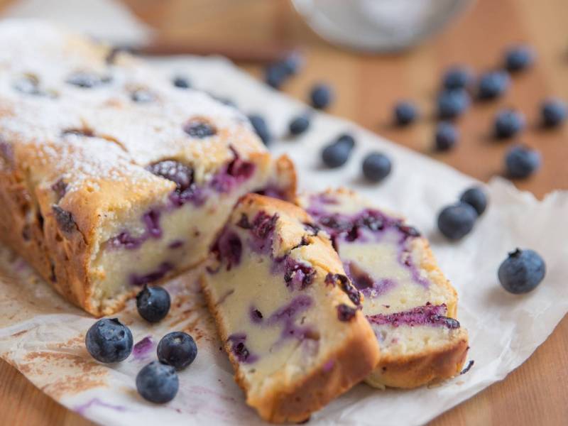 Blaubeer-Joghurt-Kuchen auf Backpapier mit zwei abgeschnittenen Stückchen.