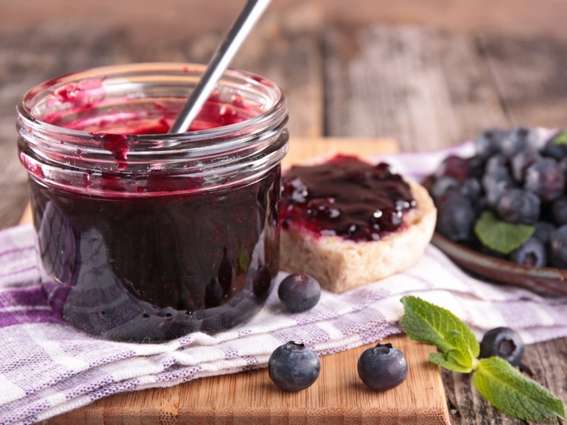 Ein offenes Glas Blaubeer-Limetten-Konfitüre, in dem ein Löffel steckt. Daneben frische Blaubeeren und ein Brot mit der Konfitüre bestrichen.