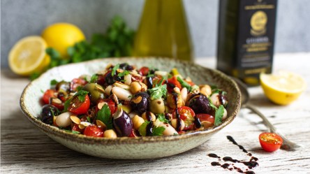 Ein Teller mit Bohnen-Oliven-Salat. Im Hintergrund liegen Zitronen, frische Kräuter und es stehen Flaschen mit Olivenöl.