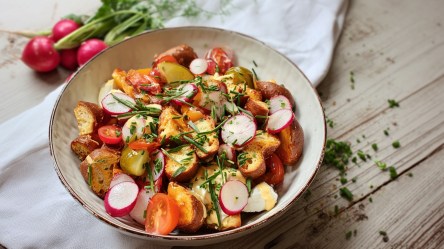Ein Teller mit Brezelsalat, der auf einem Tisch steht. Im Hintergrund kann man einen kleinen Bund Radieschen erkennen. Rechts neben dem Teller liegen frisch gehackte Kräuter verstreut.