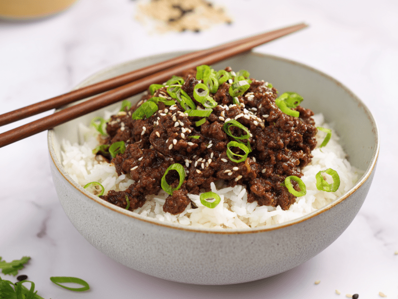 Eine Schale Bulgogi mit Hackfleisch auf hellem Untergrund. Auf der Schale liegen zwei Essstäbchen. Der Gericht ist mit Frühlingszwiebeln und Sesamsamen garniert.
