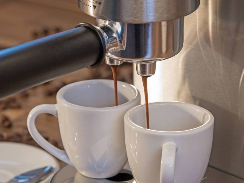 Eine Nahaufnahme, auf der ein Ausschnitt einer De'Longhi Dedica Espresso-Siebträgermaschine zu sehen ist. Es wird gerade für zwei Tassen auf einmal Espresso zubereitet. Sie stehen unter dem Siebträger, aus dem der Kaffee läuft. Im Hintergrund liegen Kaffeebohnen verstreut.