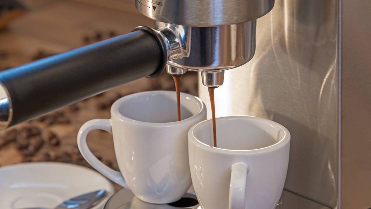 Eine Nahaufnahme, auf der ein Ausschnitt einer De'Longhi Dedica Espresso-Siebträgermaschine zu sehen ist. Es wird gerade für zwei Tassen auf einmal Espresso zubereitet. Sie stehen unter dem Siebträger, aus dem der Kaffee läuft. Im Hintergrund liegen Kaffeebohnen verstreut.