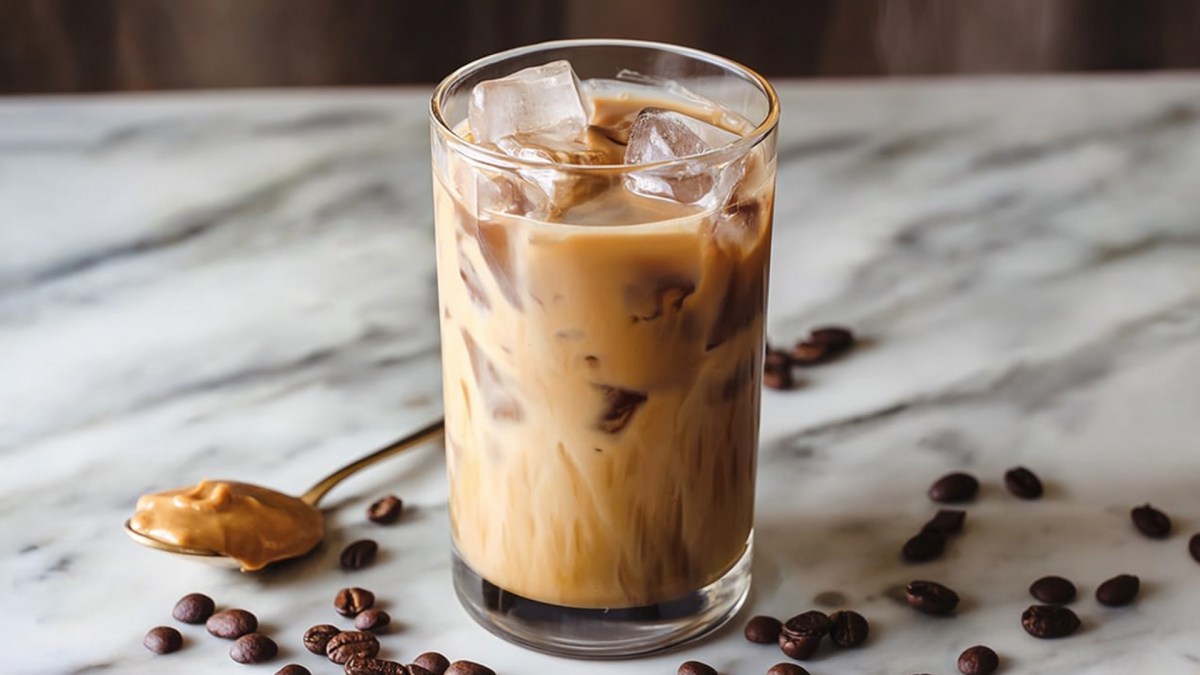 Ein Glas Erdnussbutter-Iced-Latte, das auf einer hellen marmorierten Arbeitsfläche steht. Ringsum liegen Kaffeebohnen verteilt und es ist ein Löffel voll mit Erdnussbutter zu sehen.